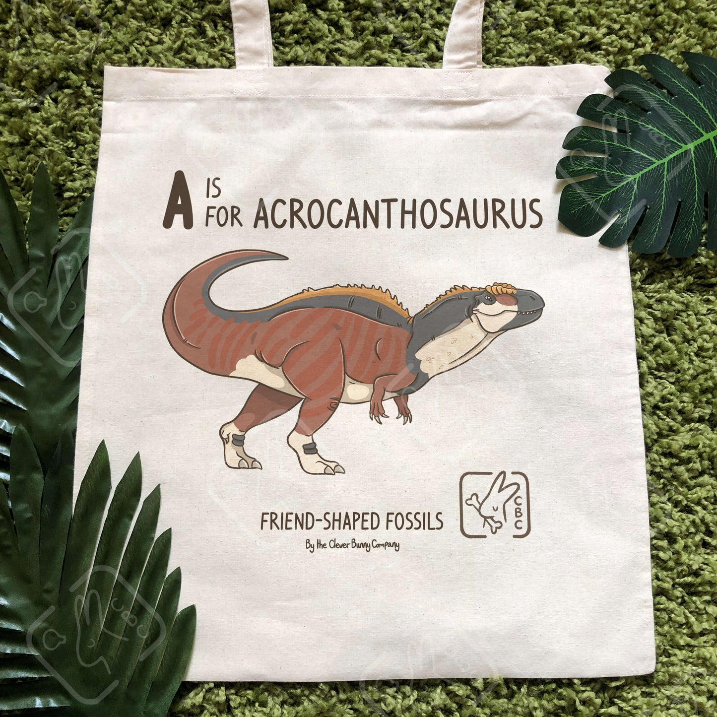 An Acrocanthosaurus printed on a cotton tote bag. The bag is on a mossy background with leaves. Text reads “A is for Acrocanthosaurus” and “Friend-Shaped Fossils by the Clever Bunny Company”.