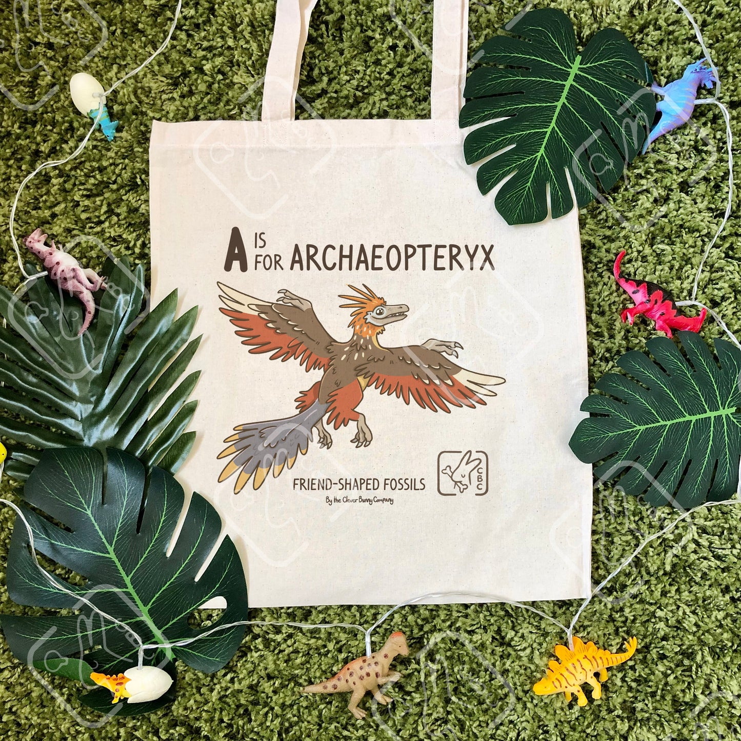 An Archaeopteryx printed on a cotton tote bag. The bag is on a mossy background with leaves. Text reads “A is for Archaeopteryx” and “Friend-Shaped Fossils by the Clever Bunny Company”.