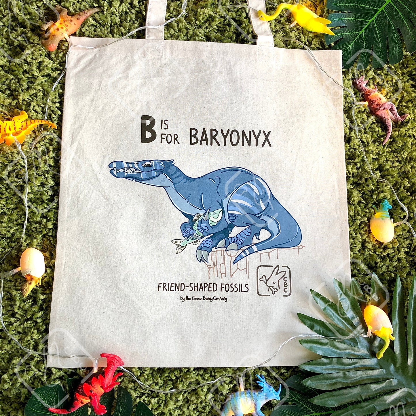 A cotton tote bag featuring a blue baryonyx dinosaur holding a fish. Text reads "B is for Baryonyx" and "Friend-shaped Fossils by the Clever Bunny Company." The bag is on a mossy background with leaves and dinosaur string lights.
