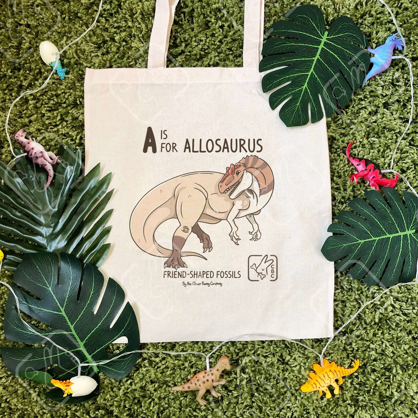 An Allosaurus printed on a cotton tote bag. The bag is on a mossy background with leaves and dinosaur string lights. Text reads “A is for Allosaurus” and “Friend-Shaped Fossils by the Clever Bunny Company”.