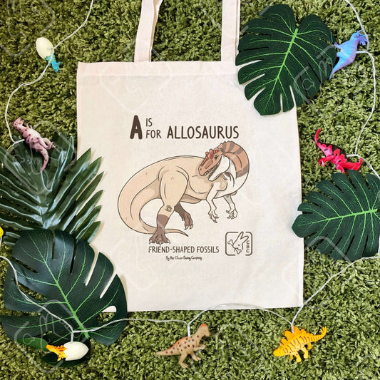 An Allosaurus printed on a cotton tote bag. The bag is on a mossy background with leaves and dinosaur string lights. Text reads “A is for Allosaurus” and “Friend-Shaped Fossils by the Clever Bunny Company”.
