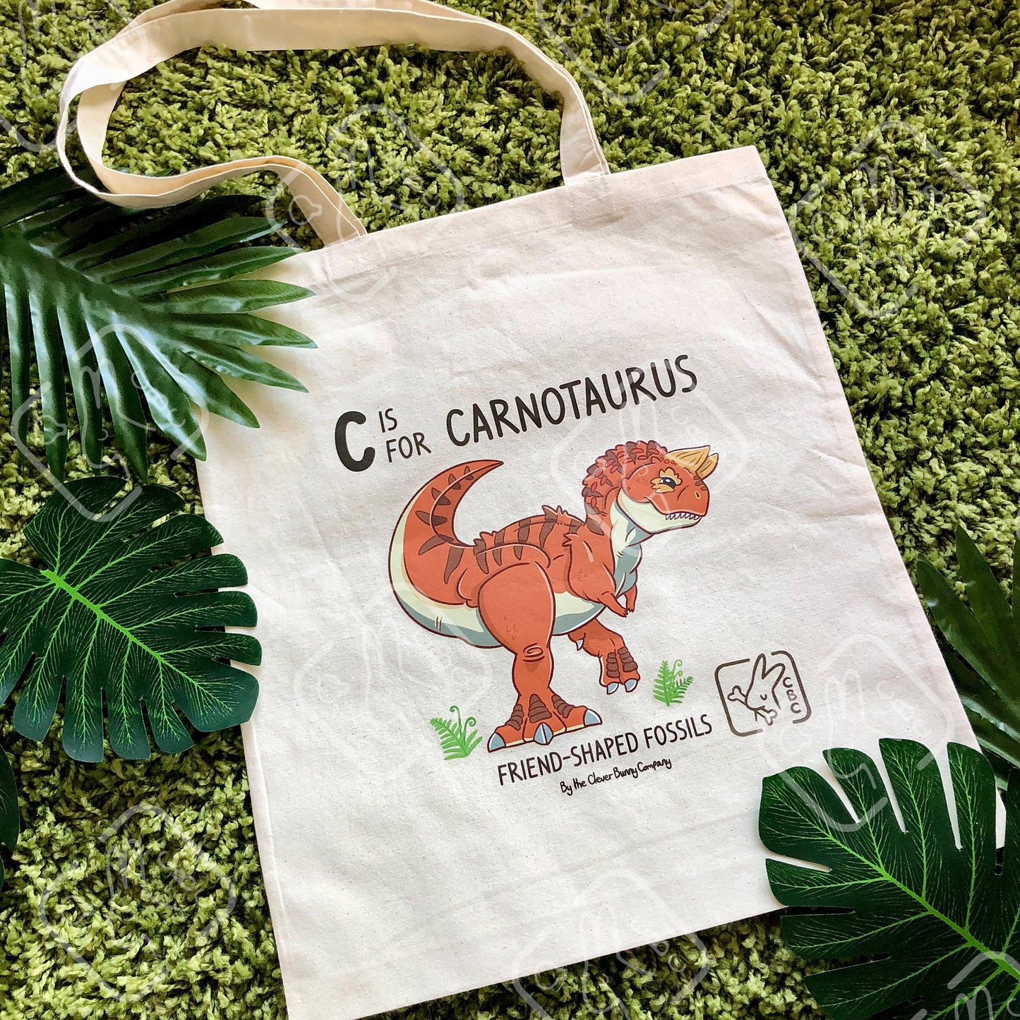 A tote bag featuring an orange Carnotaurus  dinosaur. Text reads "C is for Carnotaurus" and "Friend-shaped Fossils by the Clever Bunny Company." The bag is on a mossy background with leaves.