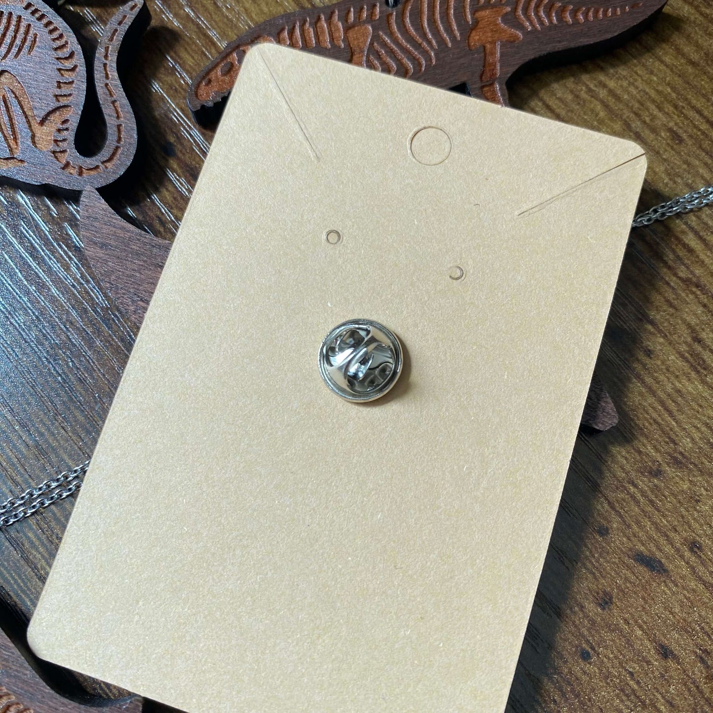 The back of the pin badge which is a small silver metal butterfly clip. It is in a Kraft paper backing card.