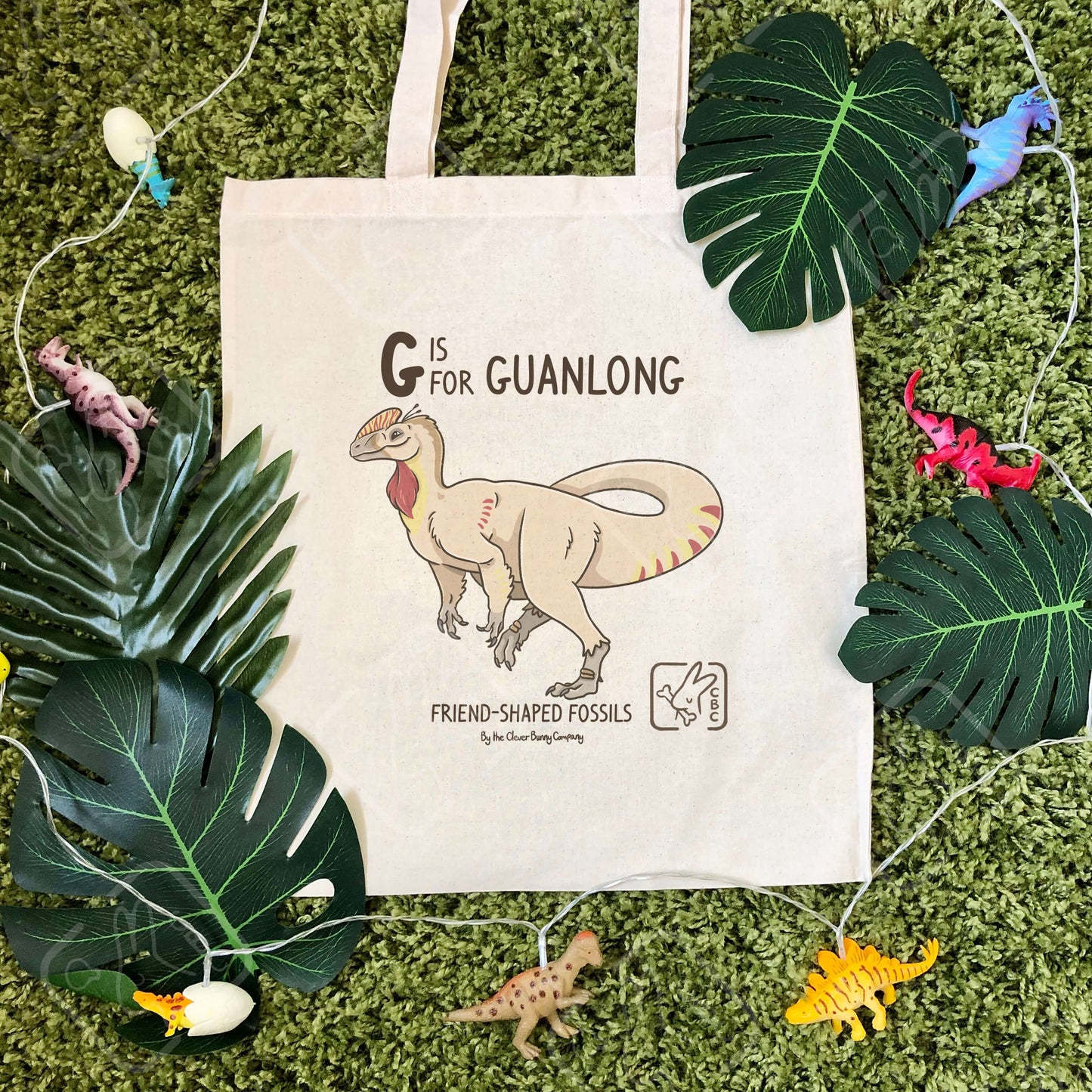 A cotton tote bag printed with a Guanlong dinosaur! Text reads “G is for Guanlong”. The tote bag is surrounded by faux leaves and dinosaur string lights.