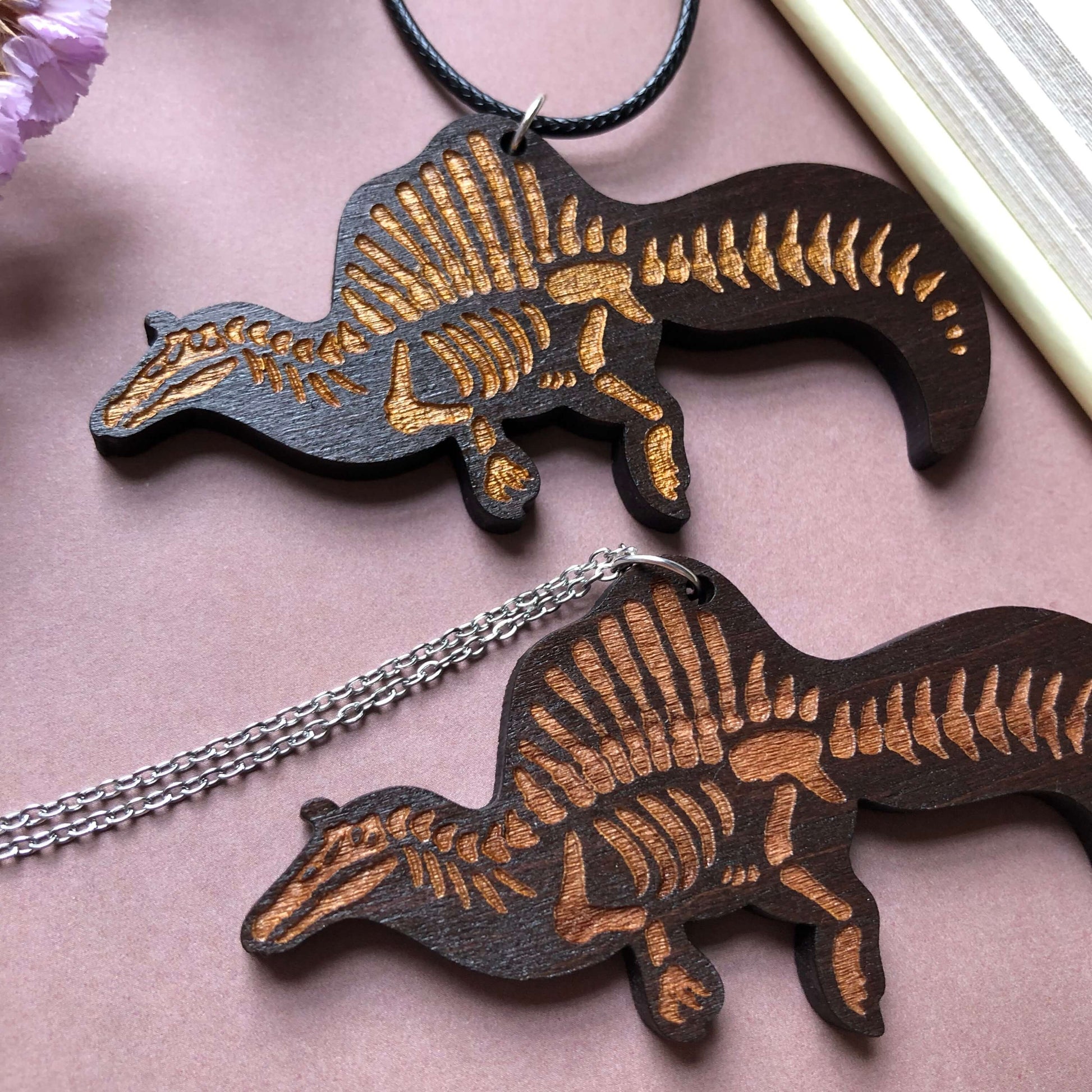 Two wooden necklaces featuring a Spinosaurus skeleton, one on a stainless steel chain and the other on a black cord. The wood is dark in colour and the skeleton on the cord is painted gold.