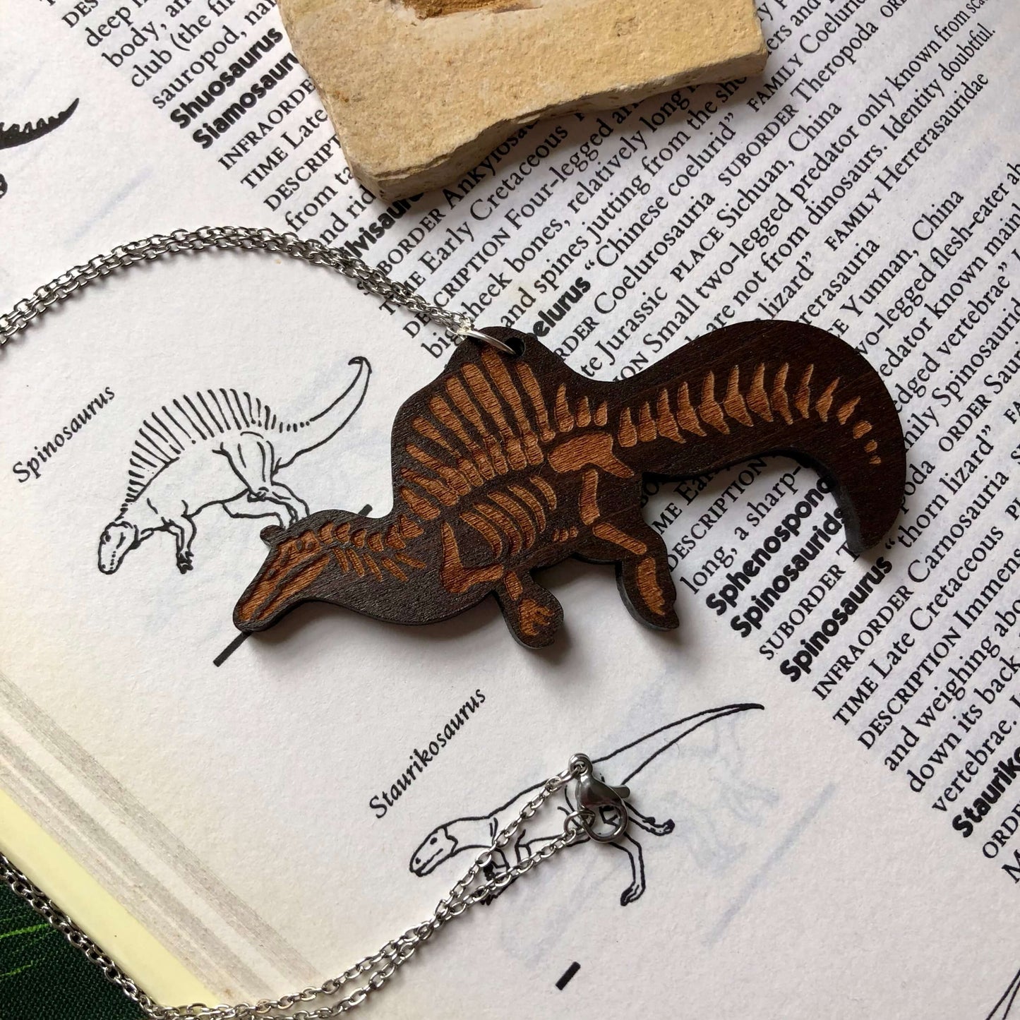 A wooden necklace featuring a Spinosaurus skeleton on a stainless steel chain. It is laying on a vintage dinosaur book. The wood is dark in colour.