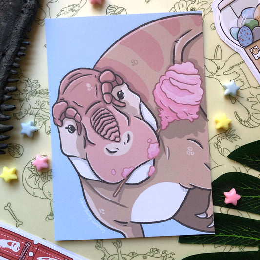 An A6 art print of a Tyrannosaurus Rex looking at the viewer and holding a stick of cotton candy in its mouth.