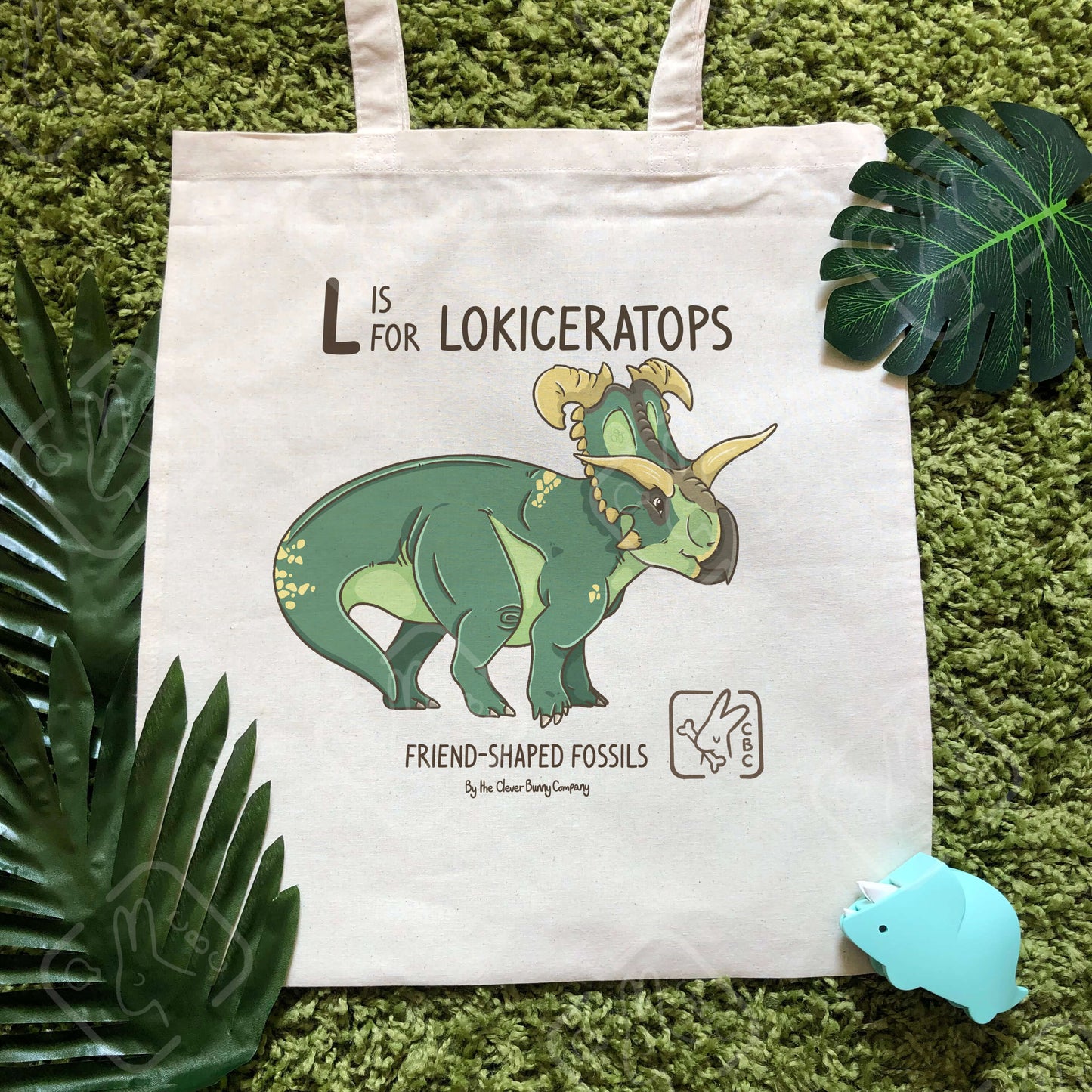 A cotton tote bag printed with a Lokiceratops dinosaur! Text reads “L is for Lokiceratops”. The tote bag is surrounded by faux leaves and a dinosaur toy.