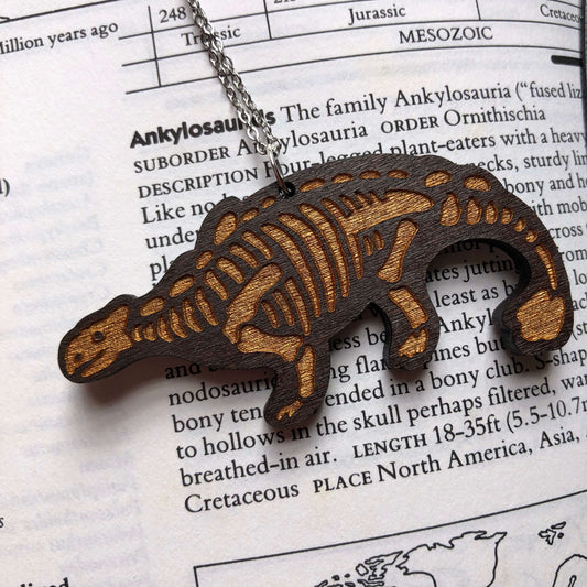 A wooden necklace of a stylised Ankylosaurus skeleton. The skeleton is engraved and then painted metallic gold. It is hanging on a stainless steel chain.
