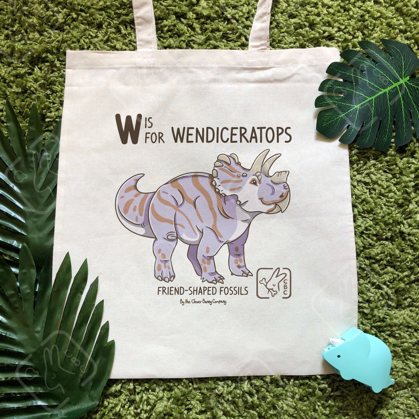 A purple and brown striped Wendiceratops dinosaur, printed on a cotton tote bag. Text reads “W is for Wendiceratops” and “Friend-Shaped Fossils by the Clever Bunny Company”. The tote bag is laying on faux moss and leaves with a ceratopsian toy beside it.