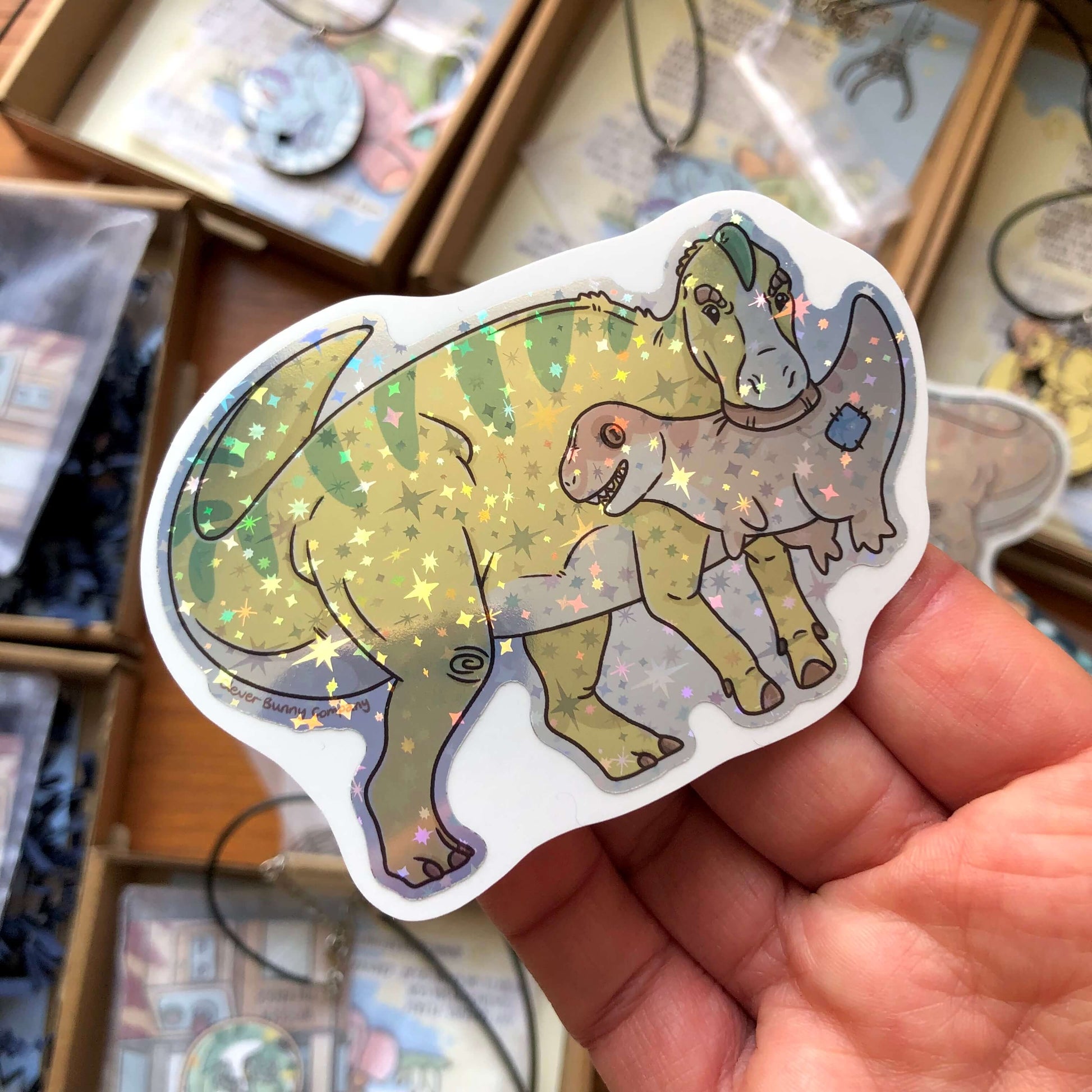 A holographic sticker of a yellow and green striped Edmontosaurus holding a Tyrannosaurus Rex plush toy in its mouth!