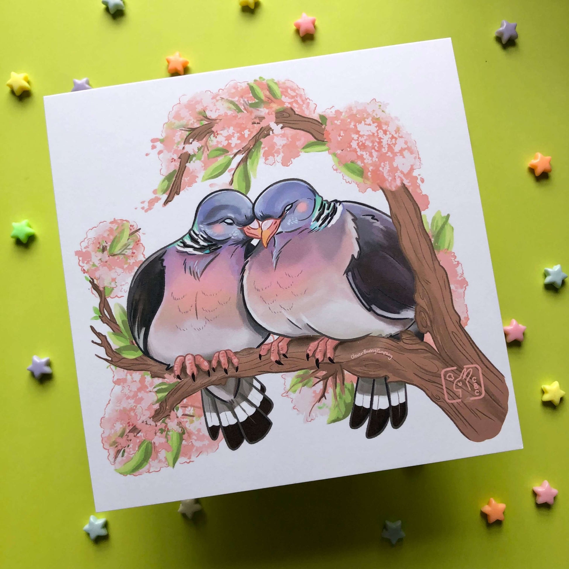 A square art print of an illustration of two woodpigeons with their heads touching. The pigeons are perched on a branch of cherry blossom.