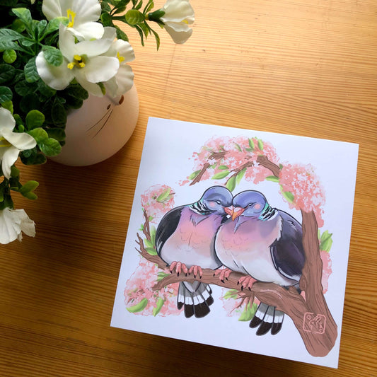A square art print of an illustration of two woodpigeons with their heads touching. The pigeons are perched on a branch of cherry blossom.