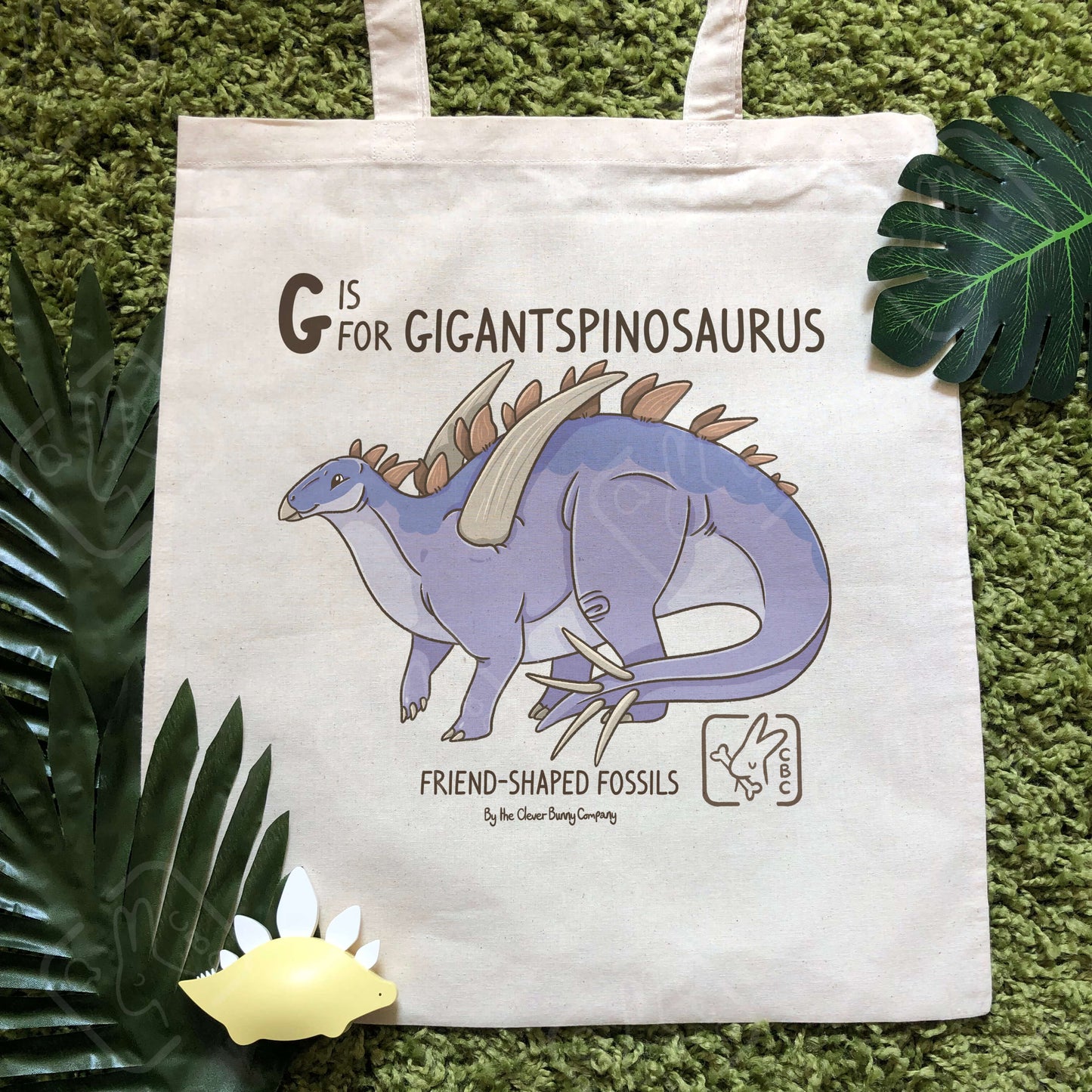 A cotton tote bag printed with a Gigantspinosaurus dinosaur! Text reads “G is for Gigantspinosaurus”. The tote bag is surrounded by faux leaves and a stegosaurus toy.