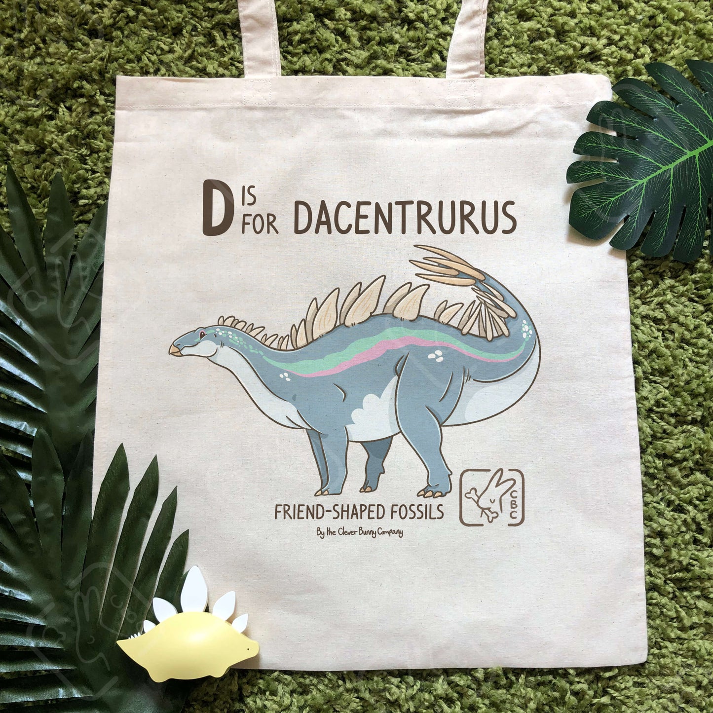 A cotton tote bag printed with a Dacentrurus dinosaur! Text reads “D is for Dacentrurus”. The tote bag is surrounded by faux leaves and a stegosaurus toy.