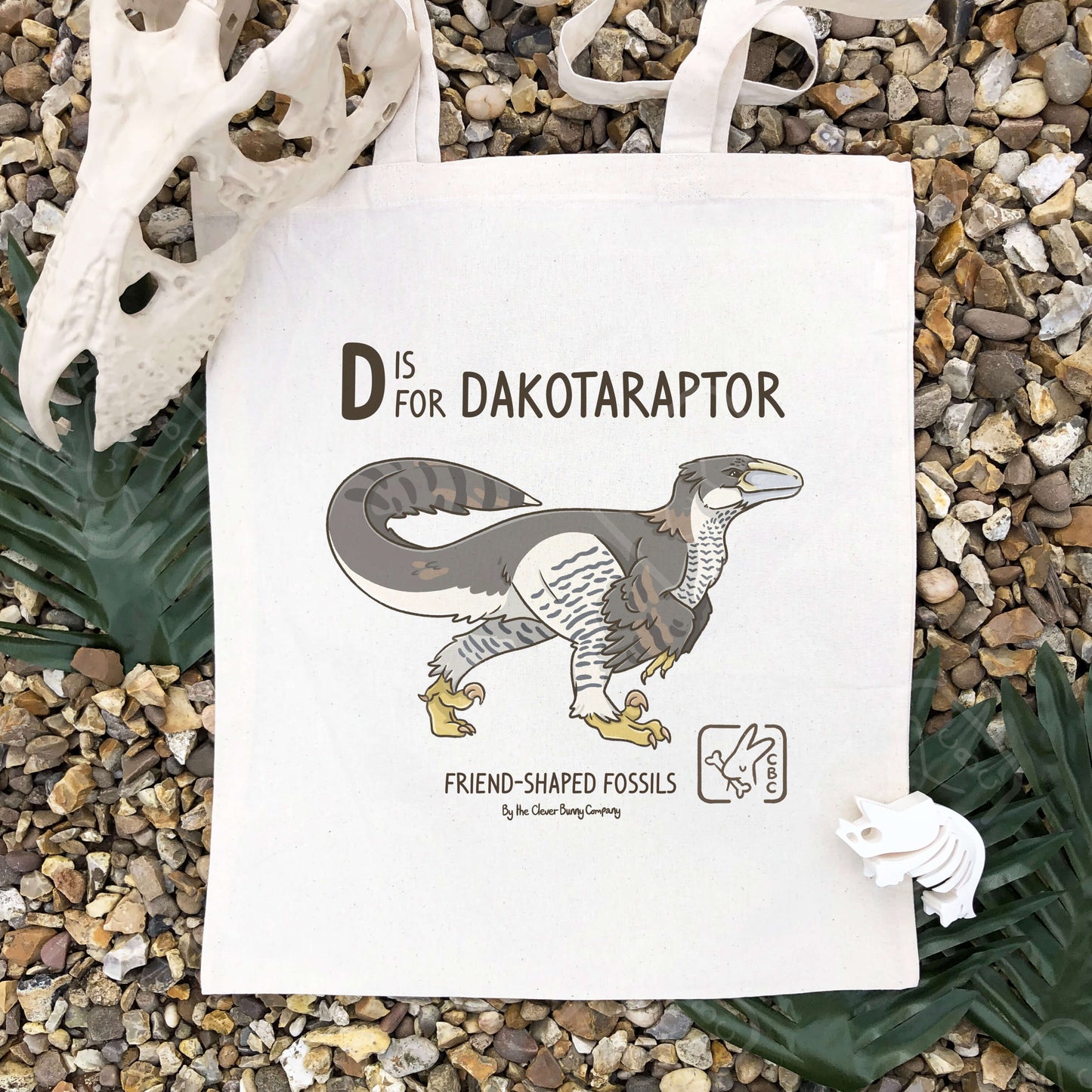 A Dakotaraptor illustration, in the colours of a Goshawk, printed on a cotton tote bag. Text reads: “D is for Dakotaraptor” and “Friend-Shaped Fossils by the Clever Bunny Company”. The bag is laying in shingle surrounded by faux leaves and dinosaur bones.