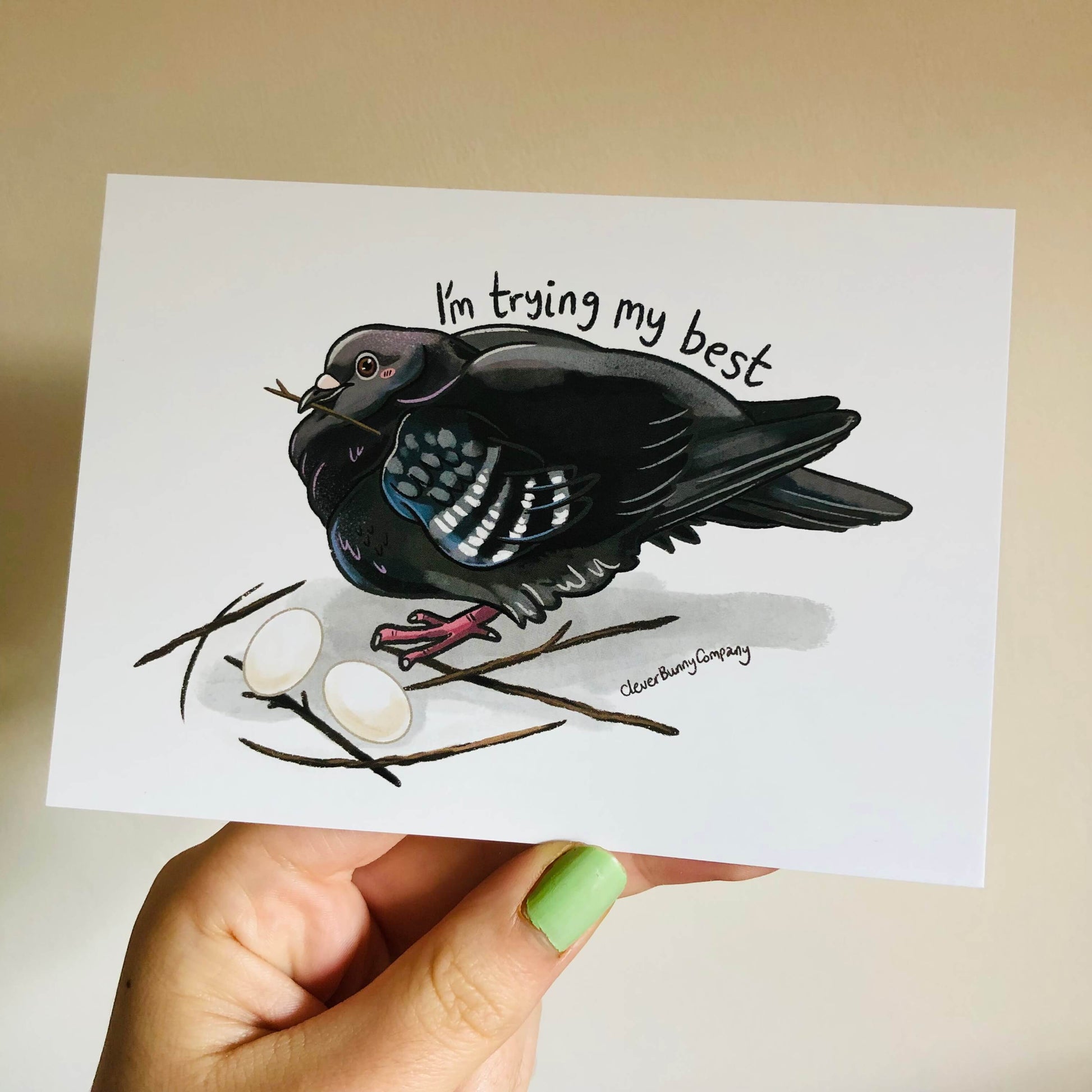 A hand holding a postcard of a pigeon with a twig in its beak sitting on a poorly built nest of 6 sticks, with two eggs. The text reads 'I'm trying my best' above the pigeon.