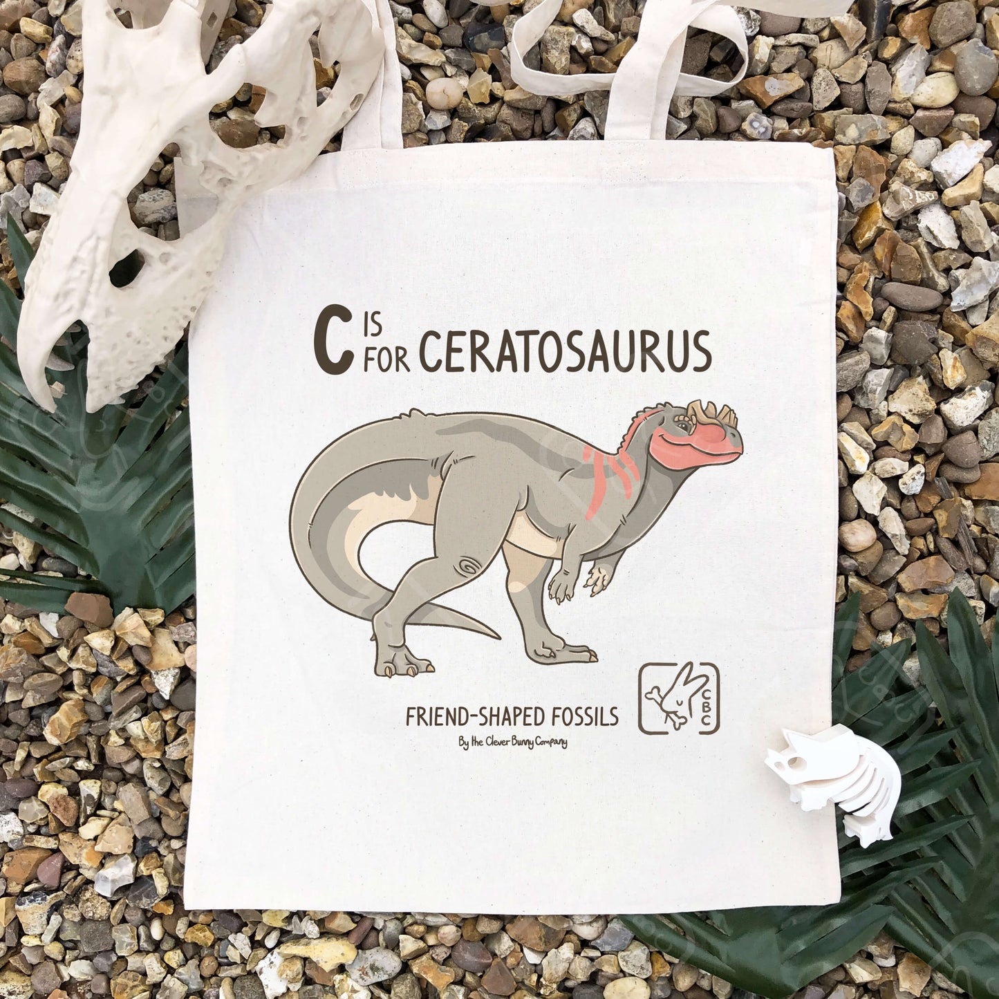 A grey, red and cream Ceratosaurus illustration, printed on a cotton tote bag. Text reads: “C is for Ceratosaurus” and “Friend-Shaped Fossils by the Clever Bunny Company”. The bag is laying in shingle surrounded by faux leaves and dinosaur bones.