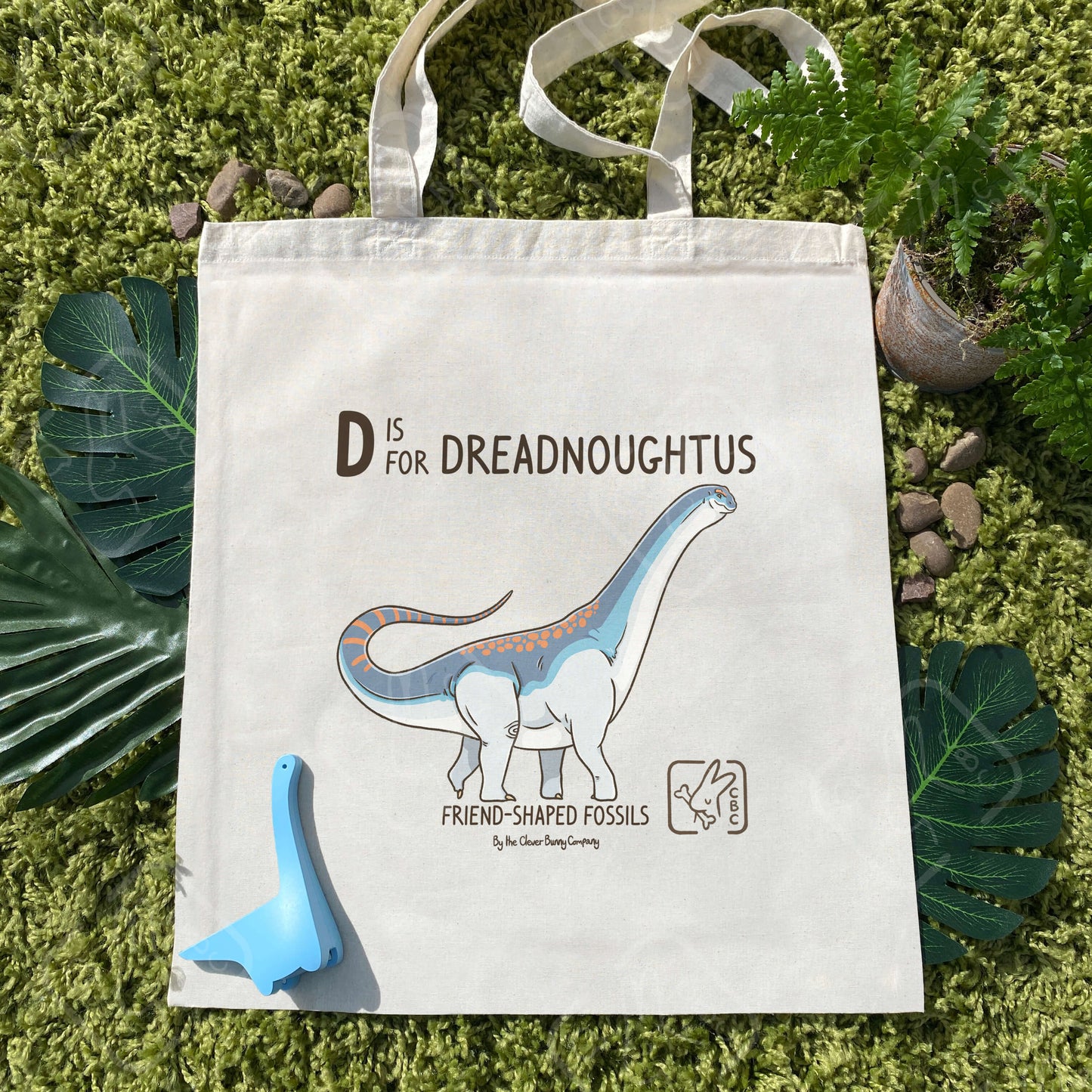 A cotton tote bag printed with a Dreadnoughtus design. It is shades of blue and white, with orange spots and stripes. Text reads: “D is for Dreadnoughtus” and “Friend-Shaped Fossils by the Clever Bunny Company.” The bag is laying on faux moss, leaves, ferns and stones, alongside a blue toy sauropod.