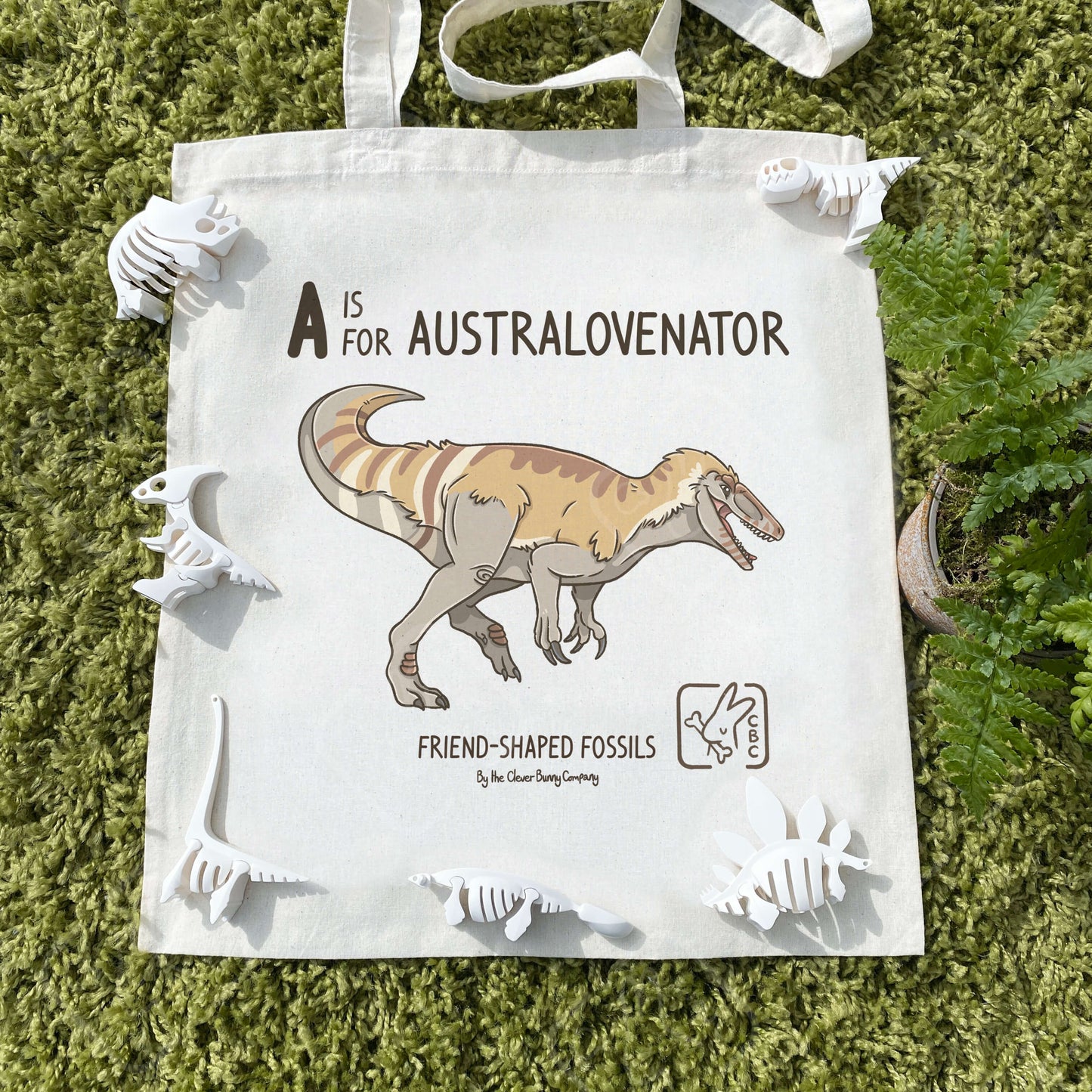 A cotton tote bag printed with an Australovenator dinosaur! Text reads “A is for Australovenator”. The tote bag is surrounded by ferns, faux plants and dinosaur skeleton toys.