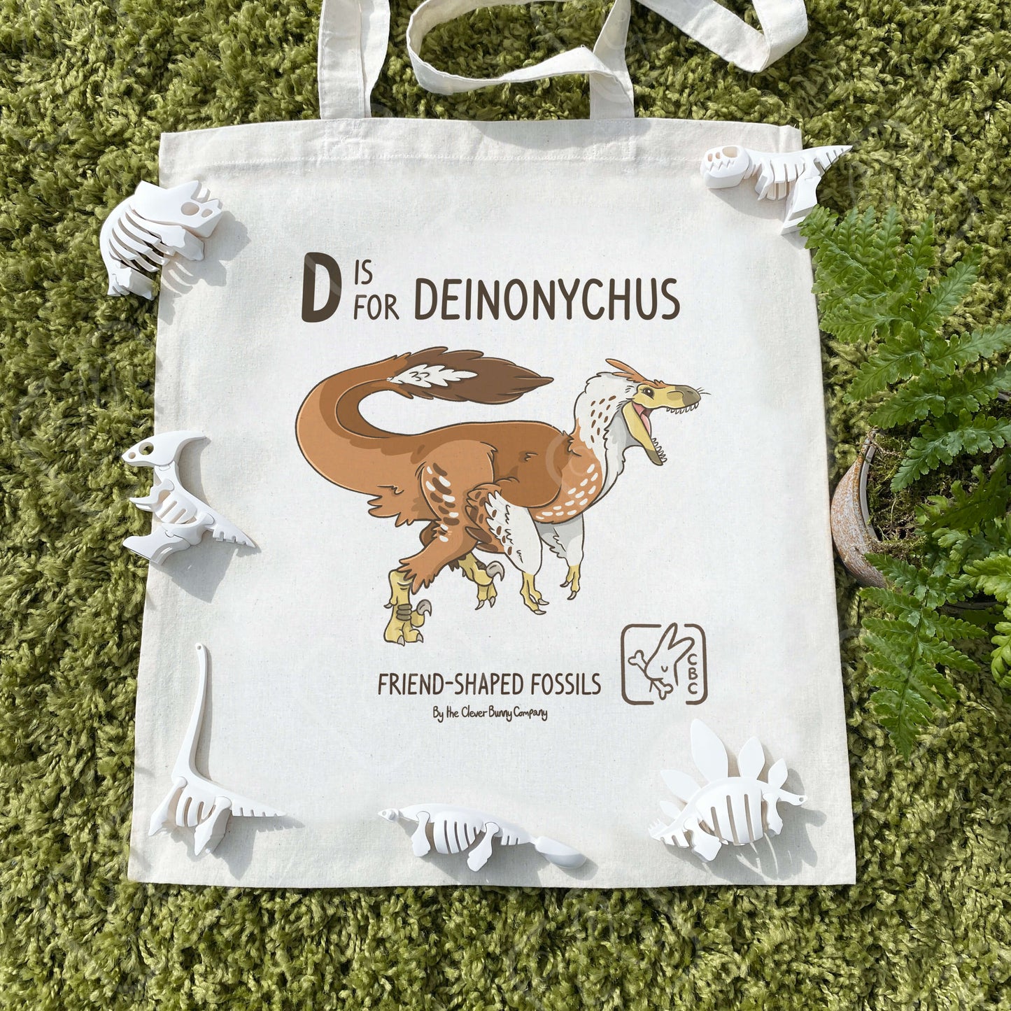 A cotton tote bag printed with a Deinonychus dinosaur! Text reads “D is for Deinonychus”. The tote bag is surrounded by faux leaves and dinosaur skeleton toys.
