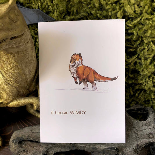 An art print of a digital illustration of a fox-coloured Nanuqsaurus. It is bracing itself against the wind, standing in the snow. Text reads “it heckin WIMDY”.