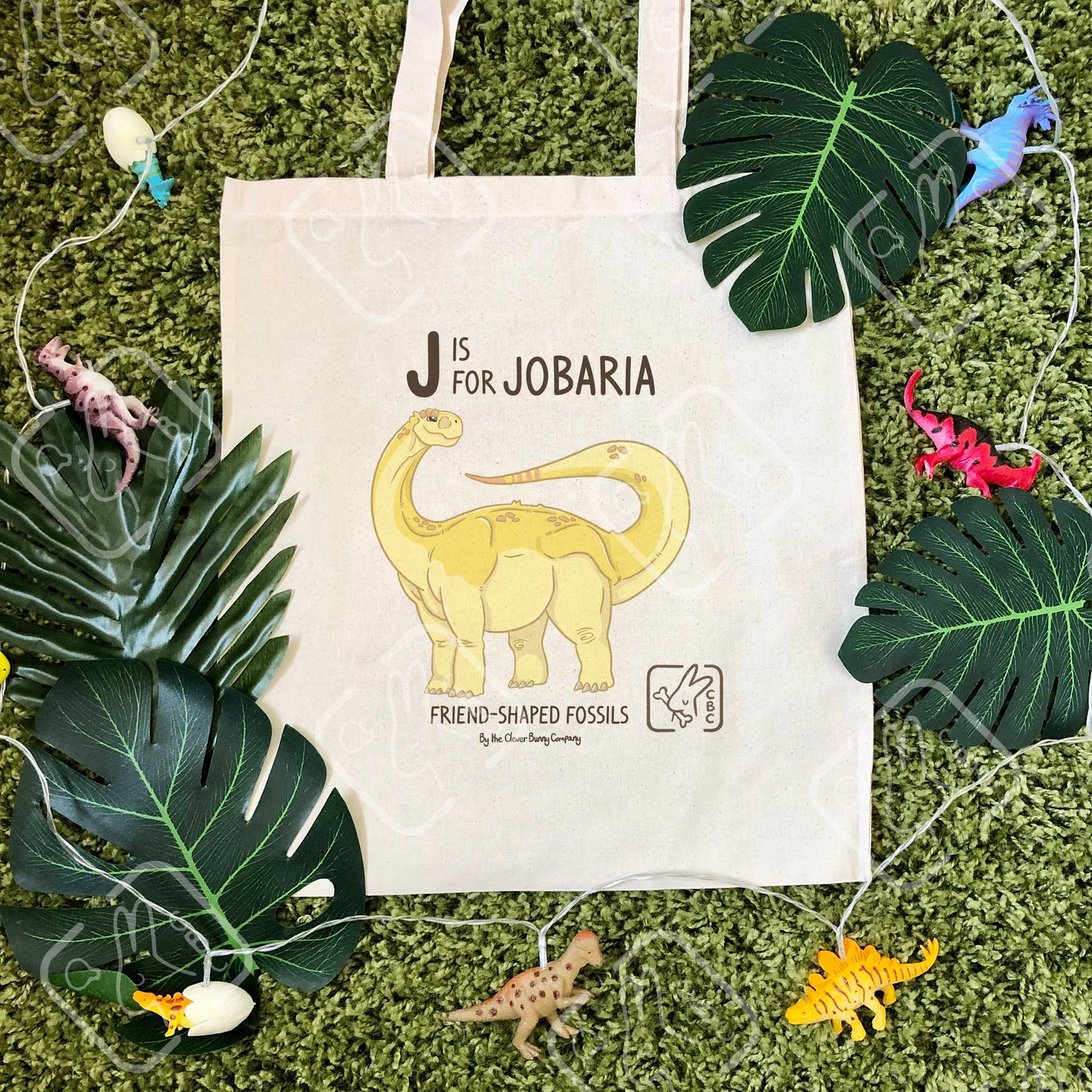 A yellow Jobaria sauropod dinosaur printed on a cotton tote bag. The bag is on a mossy background with leaves. Text reads “J is for Jobaria” and “Friend-Shaped Fossils by the Clever Bunny Company”.