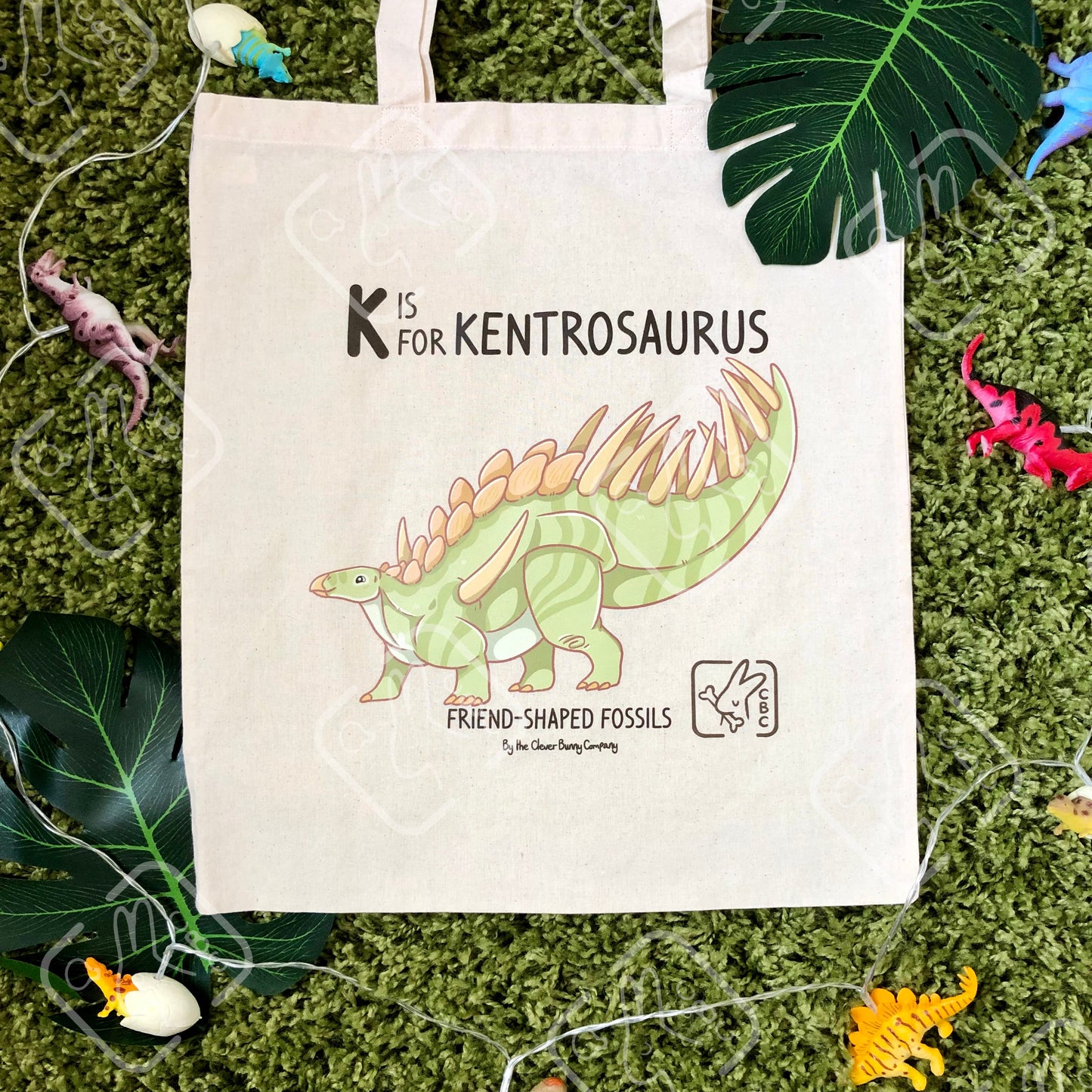 A green Kentrosaurus sauropod dinosaur printed on a cotton tote bag. The bag is on a mossy background with leaves. Text reads “K is for Kentrosaurus” and “Friend-Shaped Fossils by the Clever Bunny Company”.