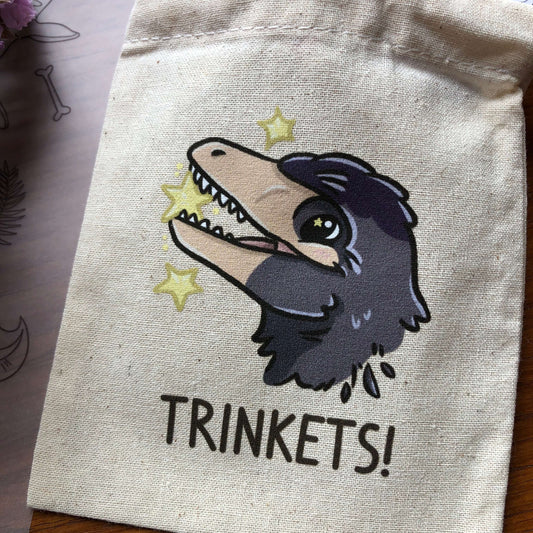 A small drawstring cotton bag printed with a Microraptor design. The Microraptor is holding a yellow star, and below it says “Trinkets!”