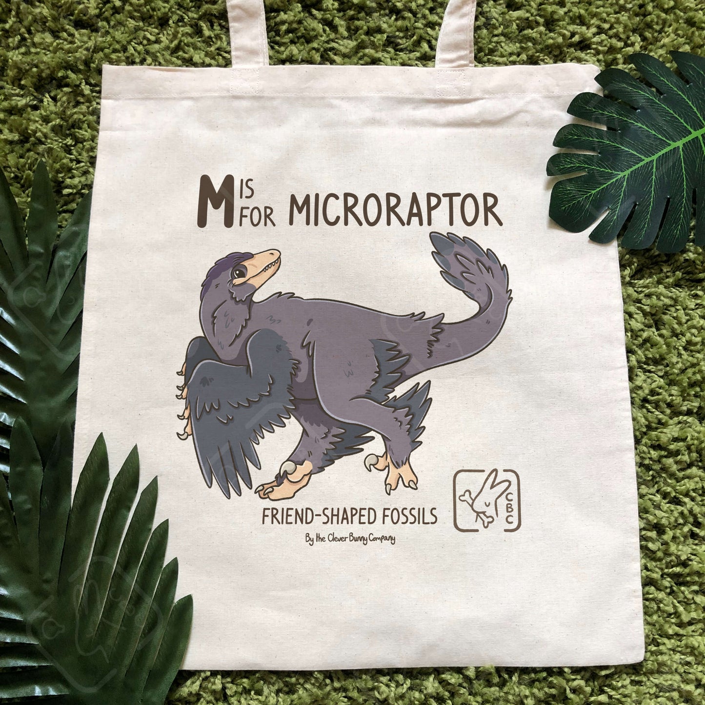 A cottons tote bag printed with a Microraptor Design! Text reads “M is for Microraptor” and “Friend-Shaped Fossils by the Clever Bunny Company”.