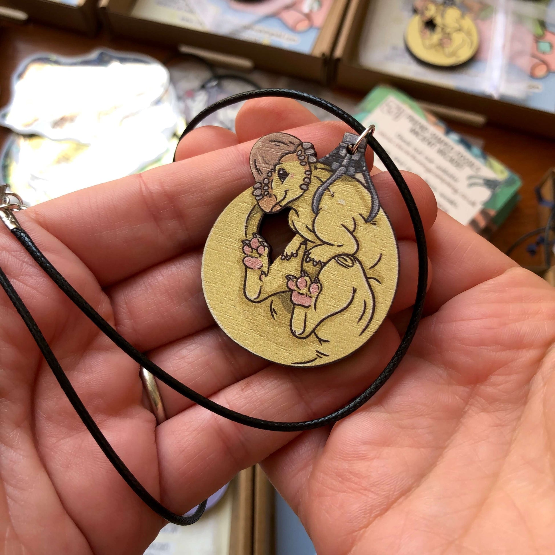 A cute yellow pachycephalosaurus necklace with pink toe beans. The necklace is made of wood with a black cotton cord.