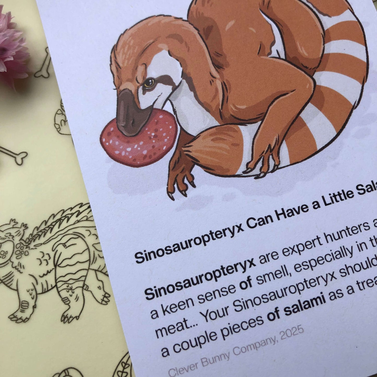 A close up of an art print of an illustration of a Sinosauropteryx dinosaur holding a slice of salami. The text below reads “Sinosauropteryx can have a little salami”. “Sinosauropteryx are expert hunters and have a keen sense of smell, especially in the case of meat…Your Sinosauropteryx should be fine with a couple pieces of salami as a treat. Clever Bunny Company, 2025.”n