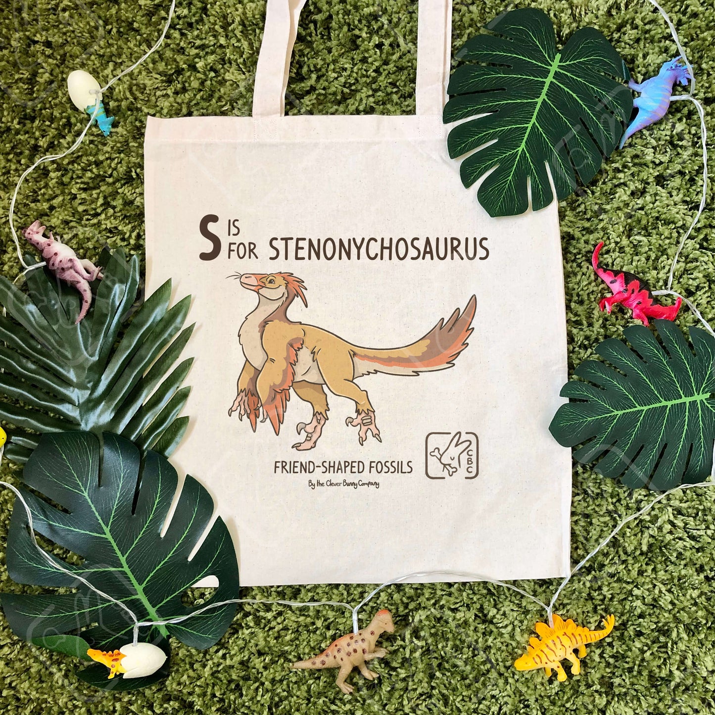 An illustration of a cute orange, red and brown Stenonychosaurus dinosaur, printed on a cotton tote bag. The bag is on faux moss, surrounded by faux leaves and dinosaur string lights.
