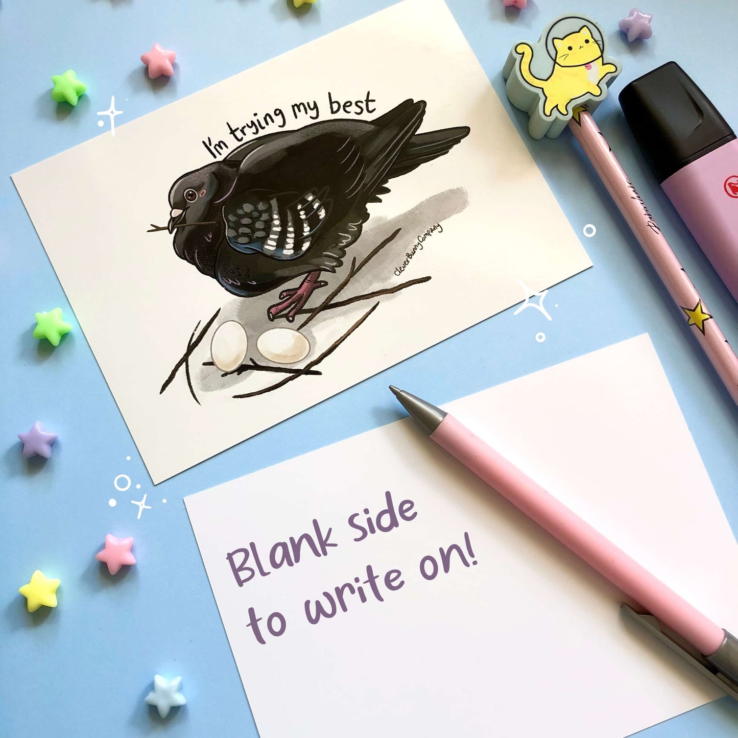 A postcard of a pigeon with a twig in its beak sitting on a poorly built nest of 6 sticks, with two eggs. The text reads 'I'm trying my best' above the pigeon. The back of the postcard is blank with the text 'Blank side to write on!'.
