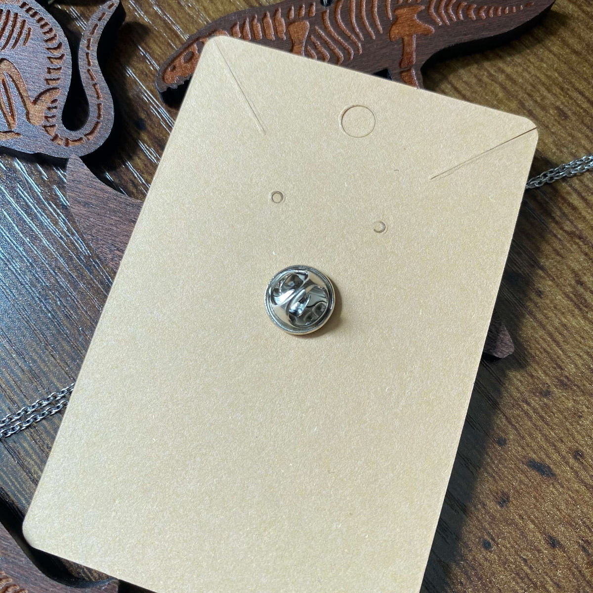The back of the pin badge which is a small silver metal butterfly clip. It is in a Kraft paper backing card.