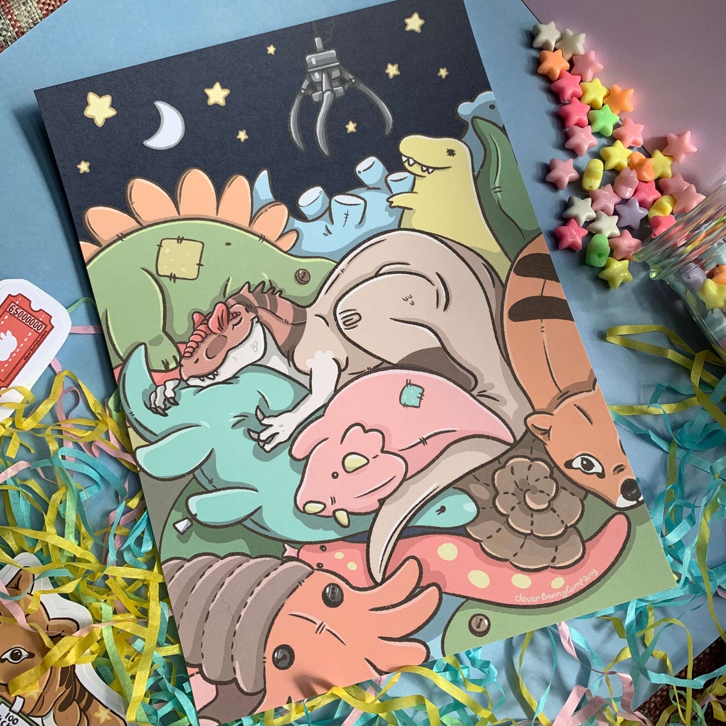 An art print of a sleepy Allosaurus, all cosy inside a claw machine full of plush prehistoric toys! There are Ancient Arcade stickers, colourful shredded paper and a glass jar of star beads around the art print as a prop.