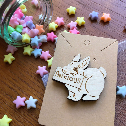 A wooden engraved pin badge shaped like a rabbit holding a sign. The sign reads “Anxious”. The badge is on a plain backing card on a table covered in colourful stars.
