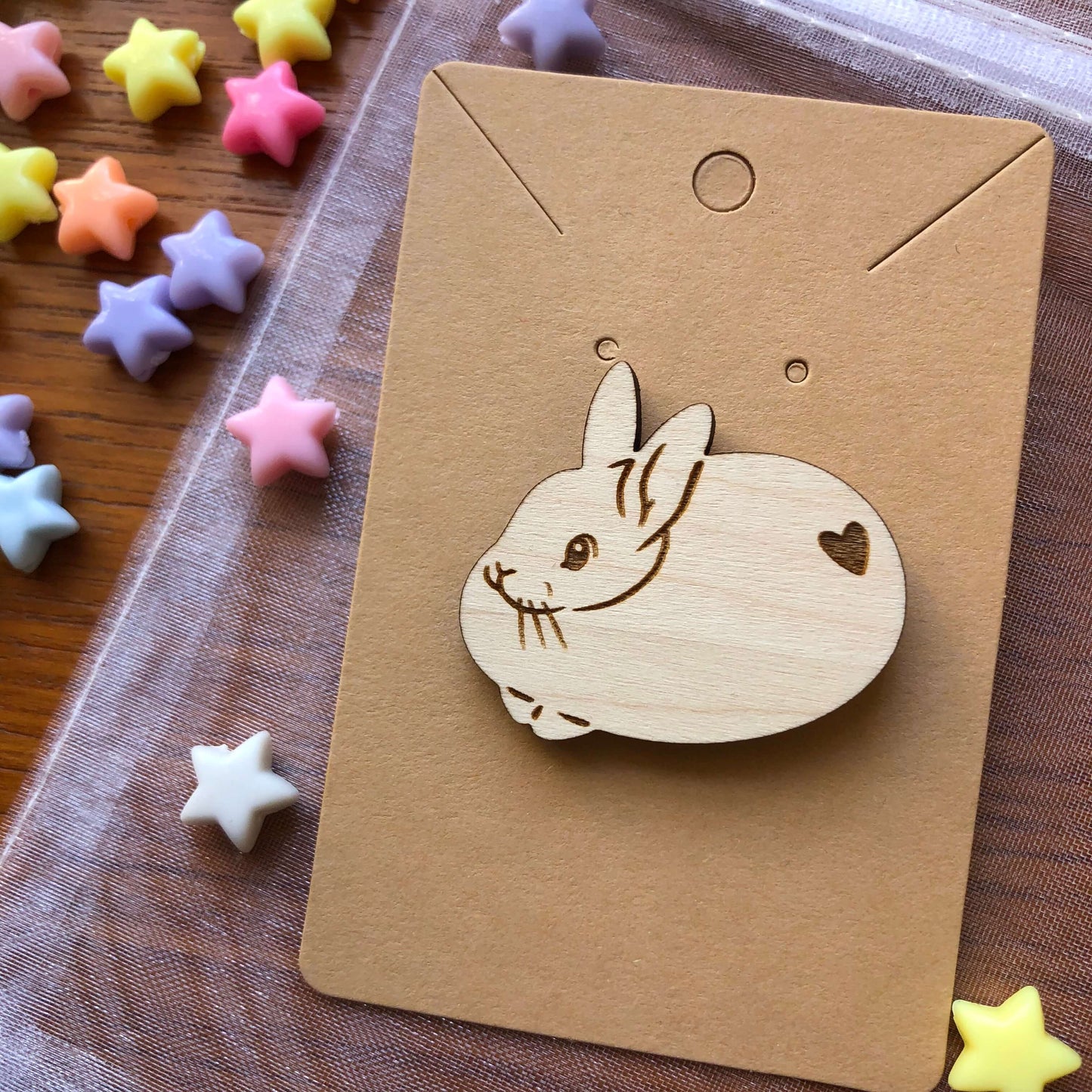 A wooden engraved pin badge shaped like a cute round rabbit with a heart shape on its bottom. The badge is on a plain backing card on an organza bag, on a table covered in colourful stars.