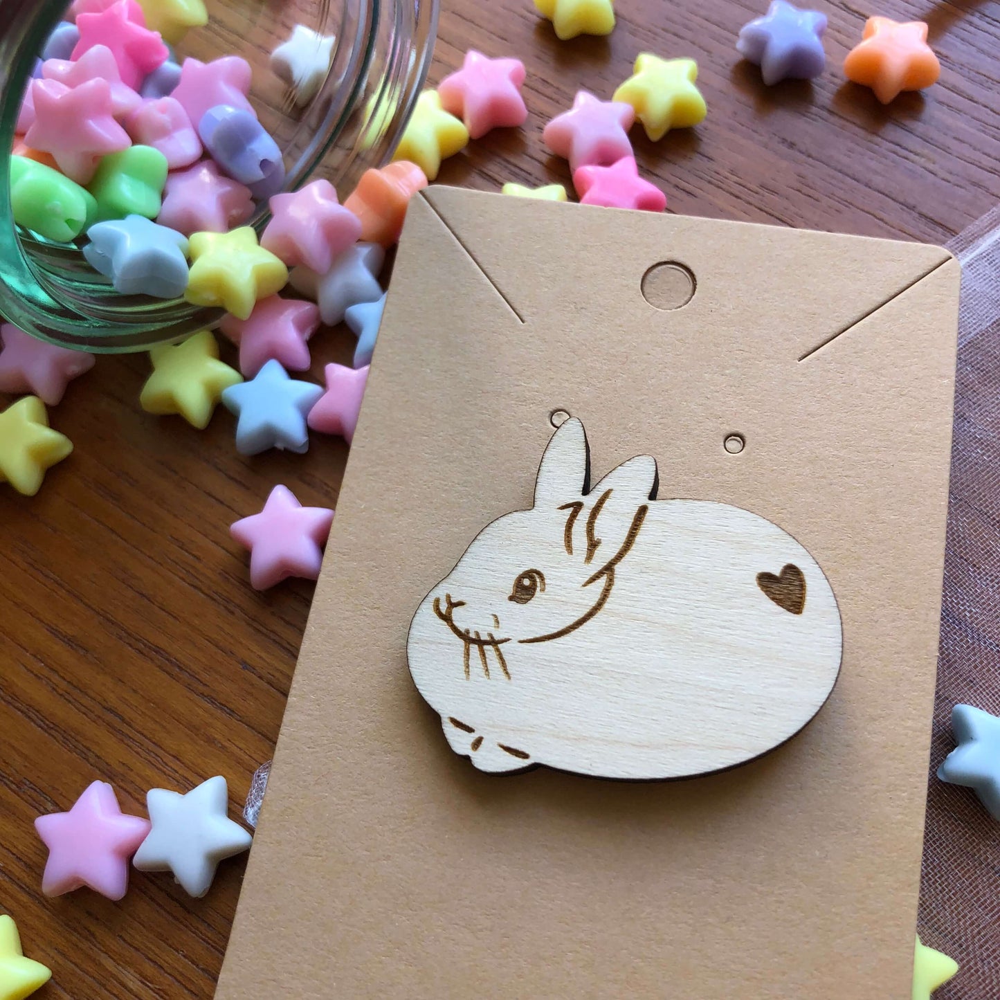 A wooden engraved pin badge shaped like a cute round rabbit with a heart shape on its bottom. The badge is on a plain backing card on a table covered in colourful stars.
