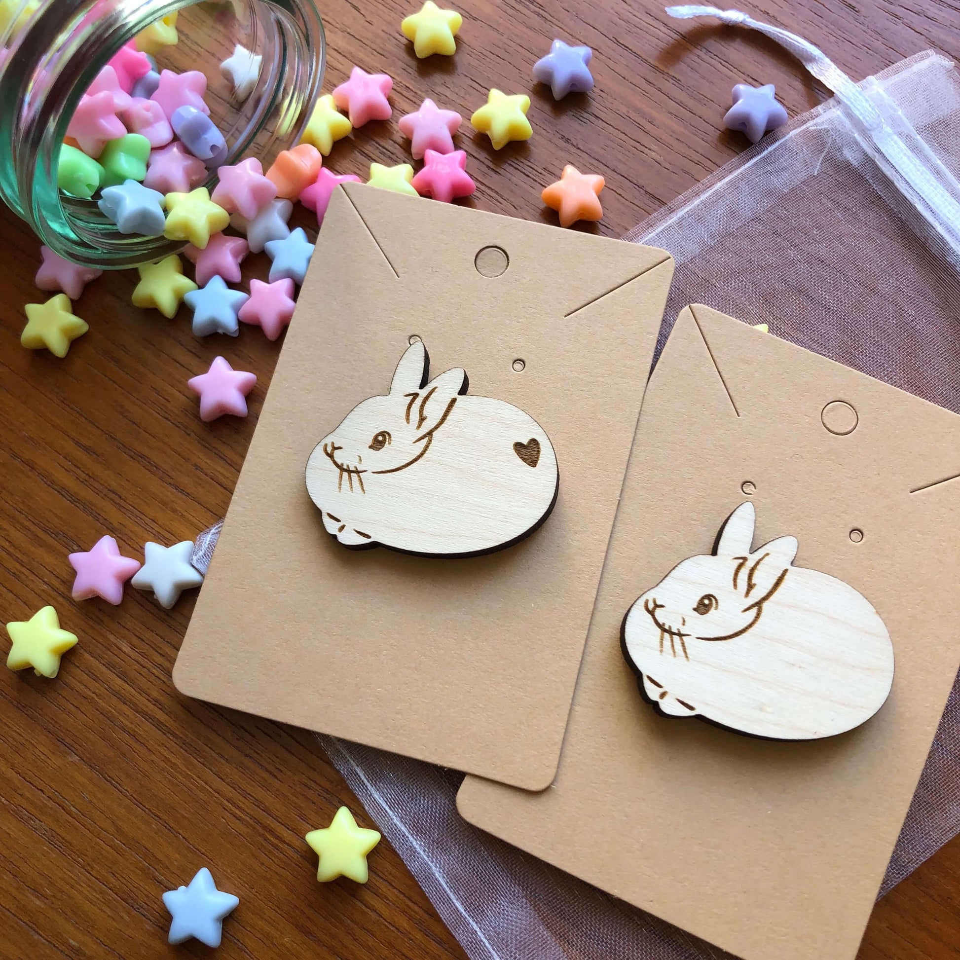 A pair of wooden engraved pin badges shaped like a cute round rabbits, one with a heart shape on its bottom and one without. The badge is on a plain backing card on a table covered in colourful stars.