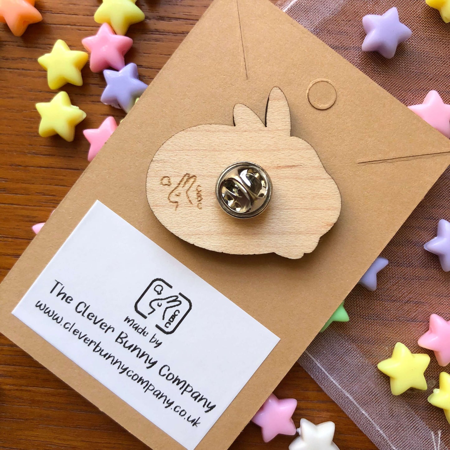 A photo showing the back of wooden engraved pin badge shaped like a rabbit. The small business logo is engraved on the back and is a rabbit with a lightbulb over the head and the letters “CBC”. The badge is on a plain backing card with the small business details printed in it, on a table covered in colourful stars.