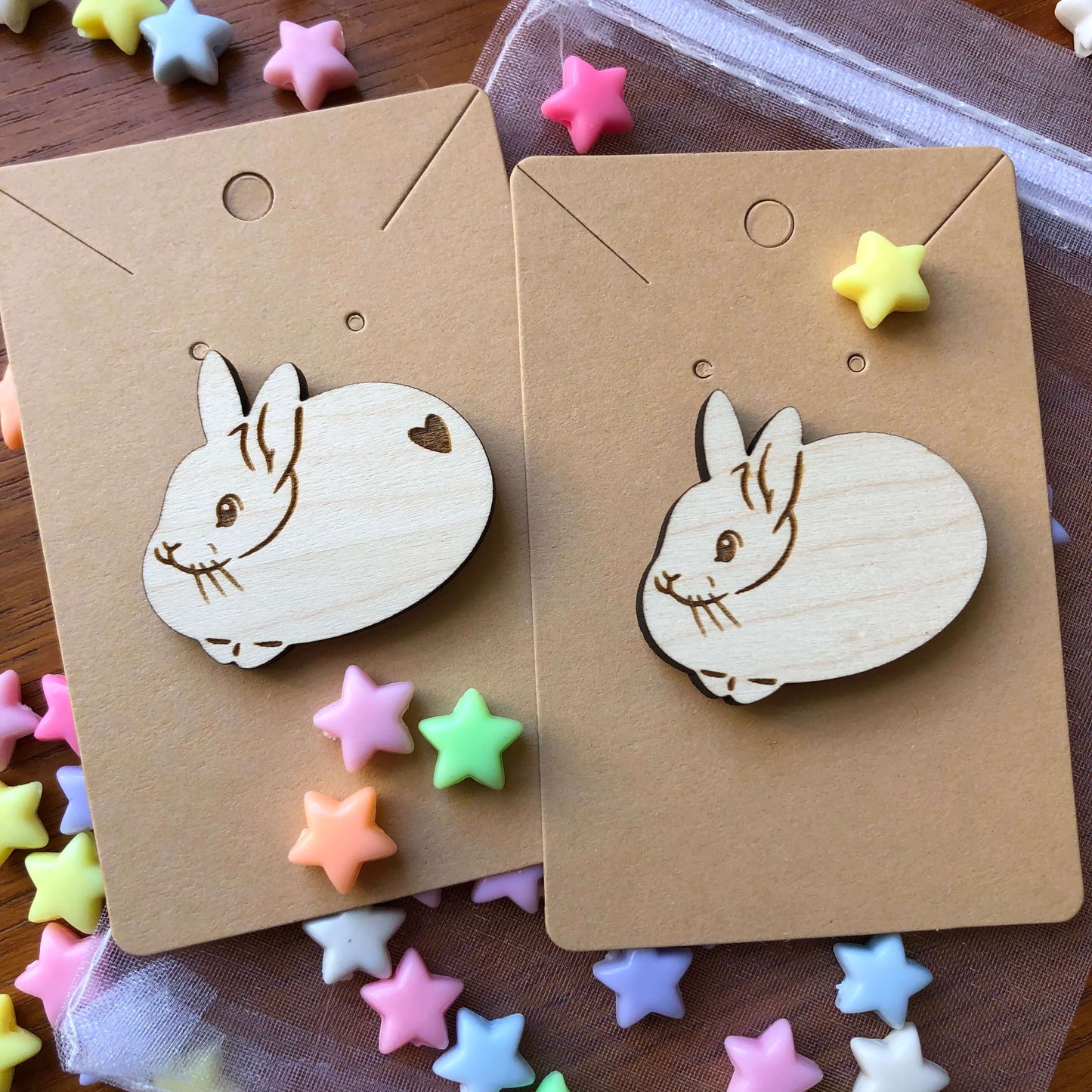 A pair of wooden engraved pin badges shaped like a cute round rabbits, one with a heart shape on its bottom and one without. The badge is on a plain backing card on a table covered in colourful stars.
