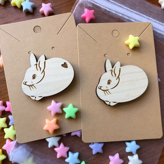A pair of wooden engraved pin badges shaped like a cute round rabbits, one with a heart shape on its bottom and one without. The badge is on a plain backing card on a table covered in colourful stars.