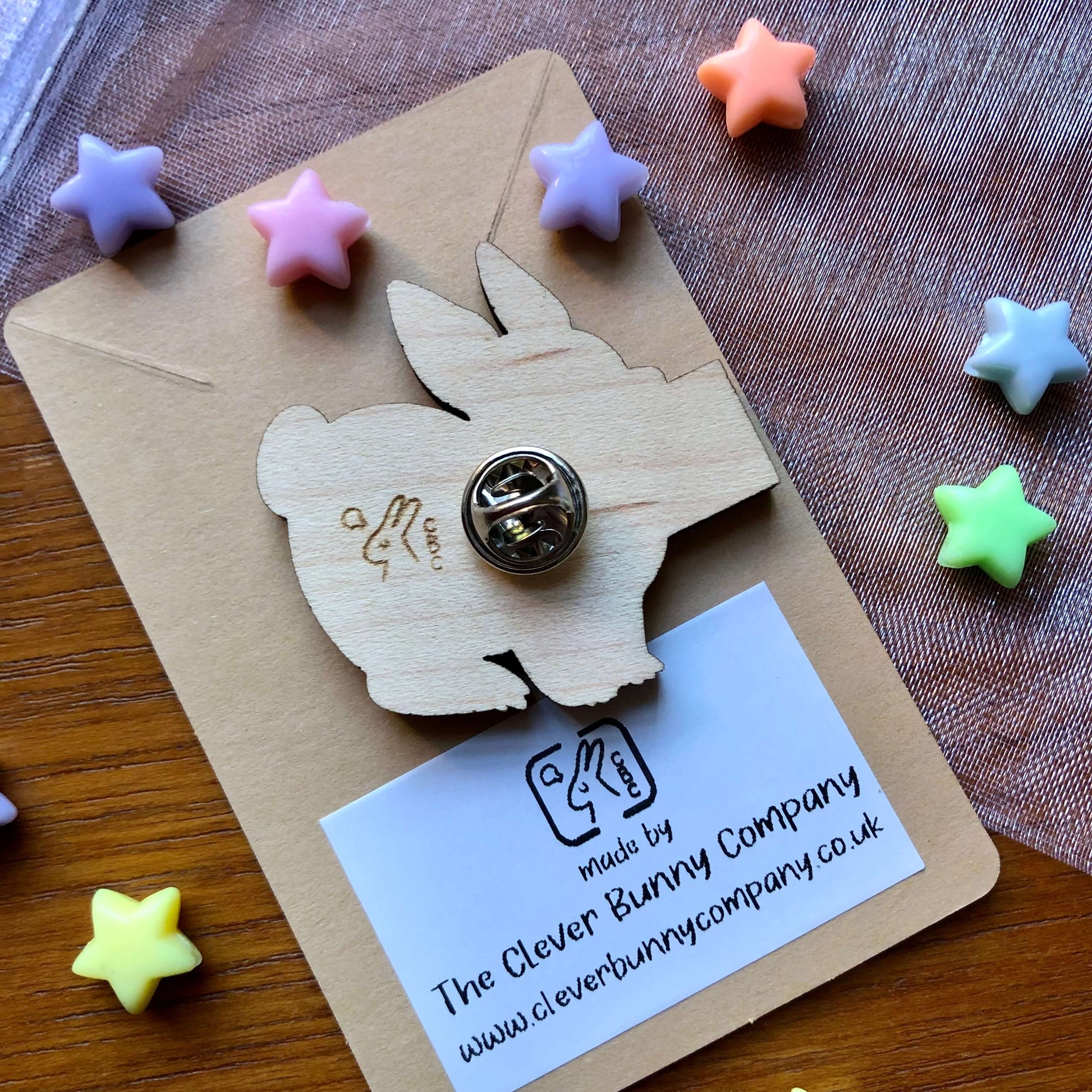 A photo showing the back of wooden engraved pin badge shaped like a rabbit holding a sign. The small business logo is engraved on the back and is a rabbit with a lightbulb over the head and the letters “CBC”. The badge is on a plain backing card with the small business details printed in it, on a table covered in colourful stars.