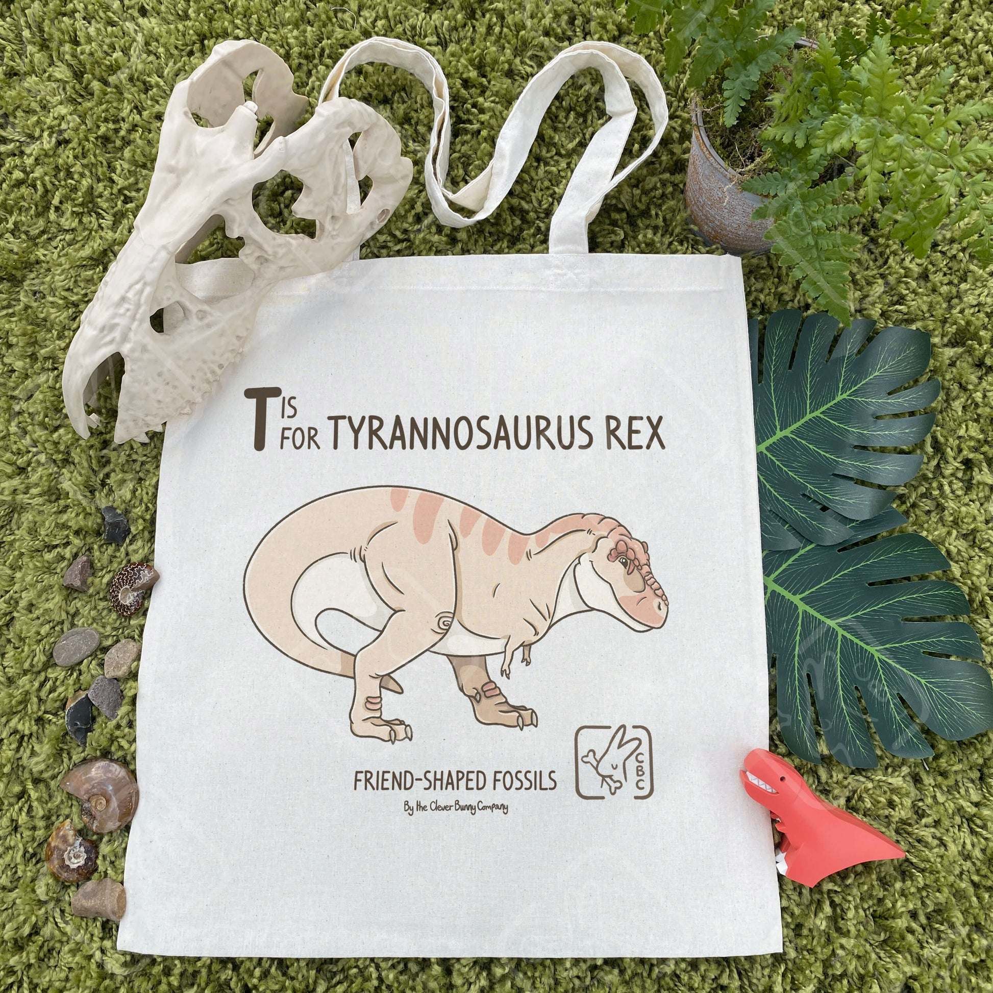 A cotton tote bag printed with a peach, striped Tyrannosaurus Rex in a more paleo-accurate style. Text reads: “T is for Tyrannosaurus Rex” and “Friend-Shaped Fossils by the Clever Bunny Company”. The background is a green mossy carpet with faux leaves, a fern, some fossils, stones and dinosaur toys.