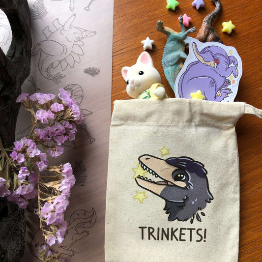 A small drawstring cotton bag printed with a Microraptor design. The Microraptor is holding a yellow star, and below it says “Trinkets!” Trinkets (like little toys and stars), are spilling out of the bag!