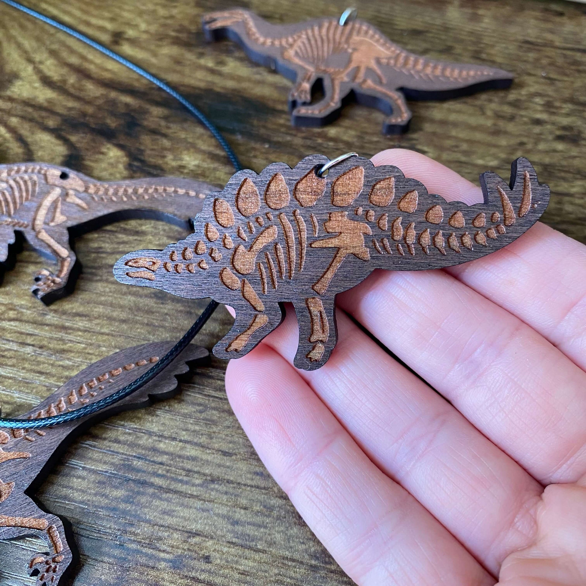 A hand holding a wooden Stegosaurus skeleton pendant, with other matching dinosaur necklaces in the background.