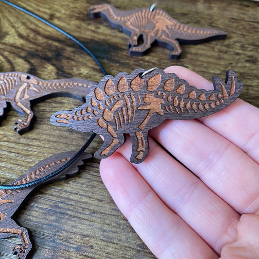 A hand holding a wooden Stegosaurus skeleton pendant, with other matching dinosaur necklaces in the background.