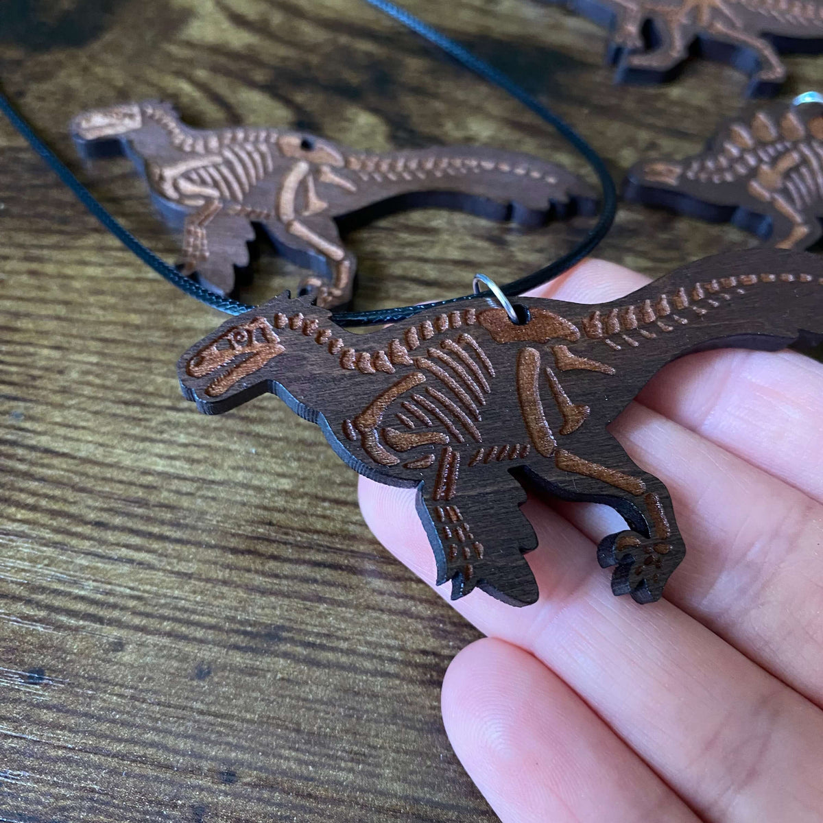 A hand holding a dark brown wooden Utahraptor skeleton necklace, with other matching wooden dinosaur skeleton pendants on black cords in the background.