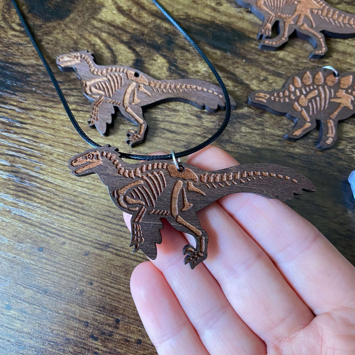 A wooden Utahraptor skeleton necklace held in a person's hand, with additional matching wooden dinosaur skeleton pendants on a black cord in the background.
